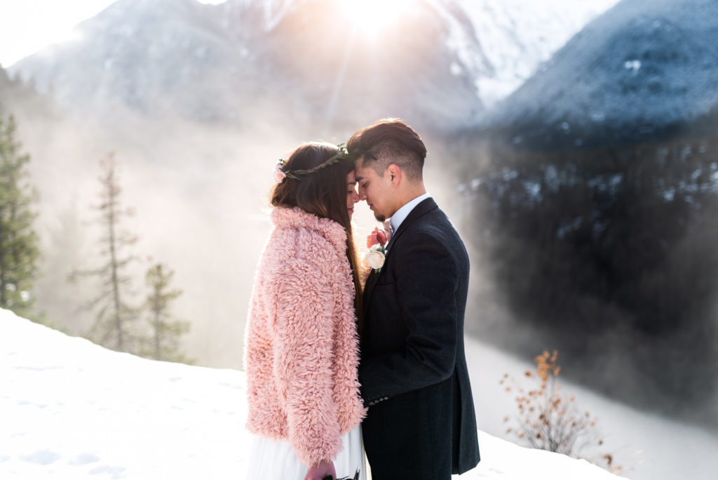 groom and bride in pink jacket standing close on snowy mountain 