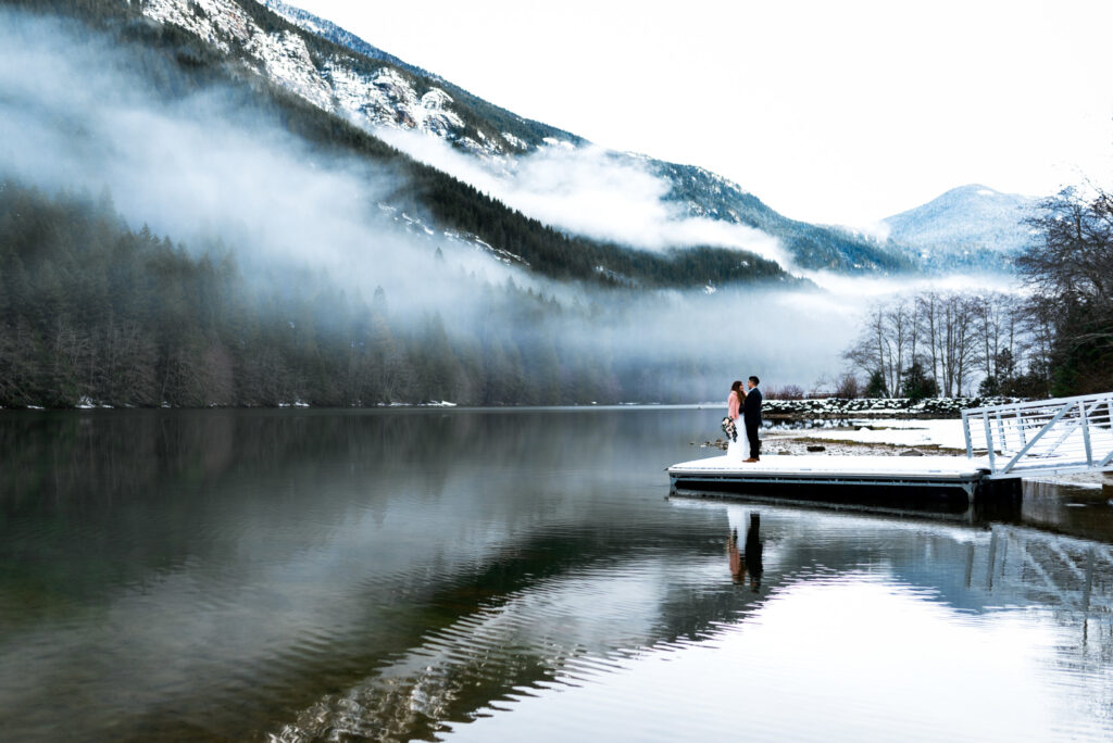 couple in wedding attire stand on dock at lake surrounded by snowy mountains