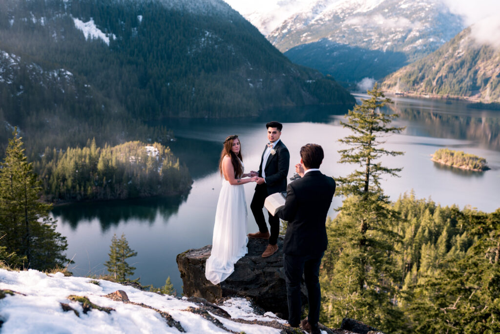 officiant and couple standing on cliffside over lake at winter elopement
