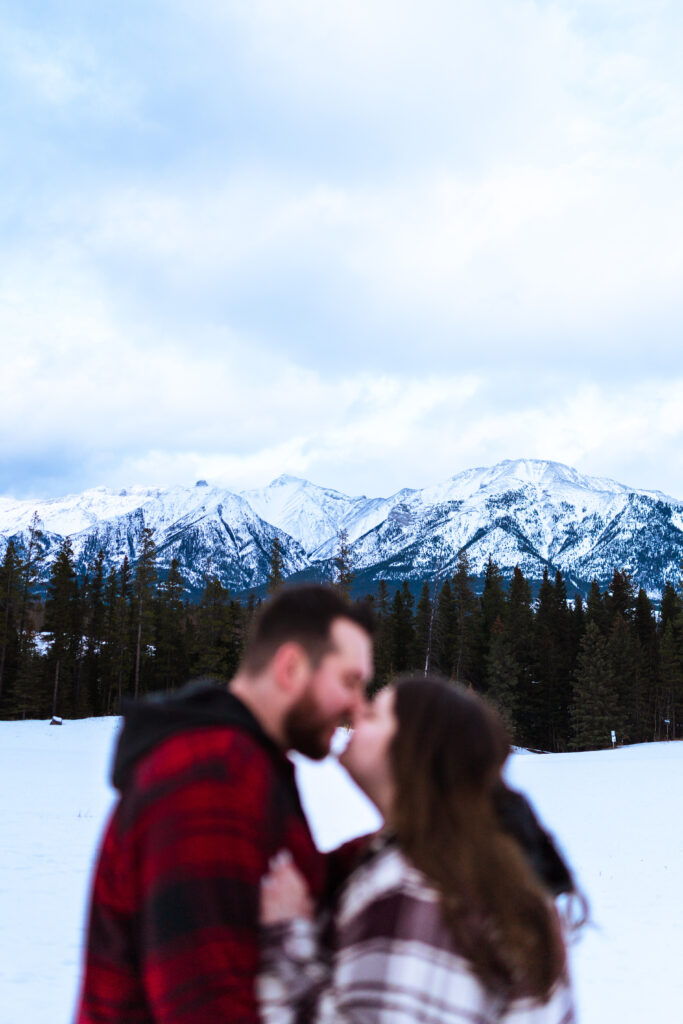 couple lean in to kiss with snow-capped Canadian mountains behind them
